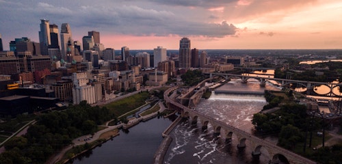 Aerial view of a city skyline at sunset with skyscrapers, a river flowing through, bridges connecting areas, and vibrant clouds above reflecting the dying light.