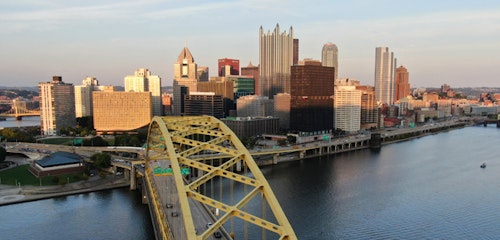 Aerial view of Pittsburgh skyline at dusk, showcasing the distinctive yellow Fort Duquesne Bridge over the Allegheny River with the city's skyscrapers in the background.