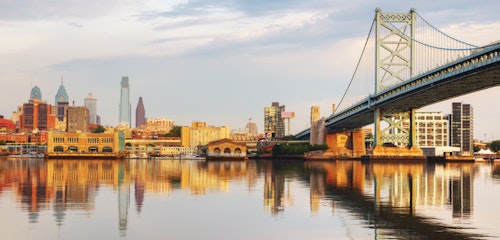 Sunrise over Philadelphia showcasing the Benjamin Franklin Bridge with a reflection on the water and the city skyline, highlighting notable skyscrapers.