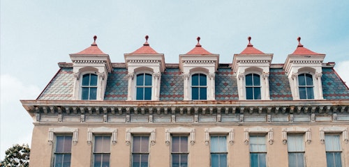 Vintage building façade with ornate details and multiple red-roofed dormers, set against a sky with light cloud cover, showcasing architectural heritage.