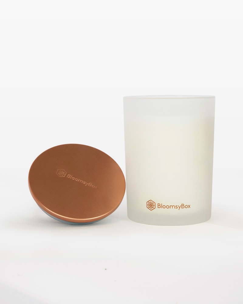 Frosted glass candle with BloomsyBox logo next to its copper-colored metal lid on a white background, reflecting a simple and elegant design.
