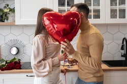 A couple, partially obscured behind a red heart-shaped balloon, shares an intimate moment in a kitchen adorned with roses, hinting at a romantic celebration.