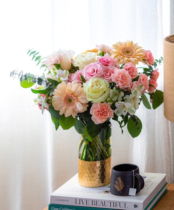 A vibrant bouquet of flowers with roses and daisies in a glass vase on a table beside stacked books with 'Live Beautiful' cover and a black mug.