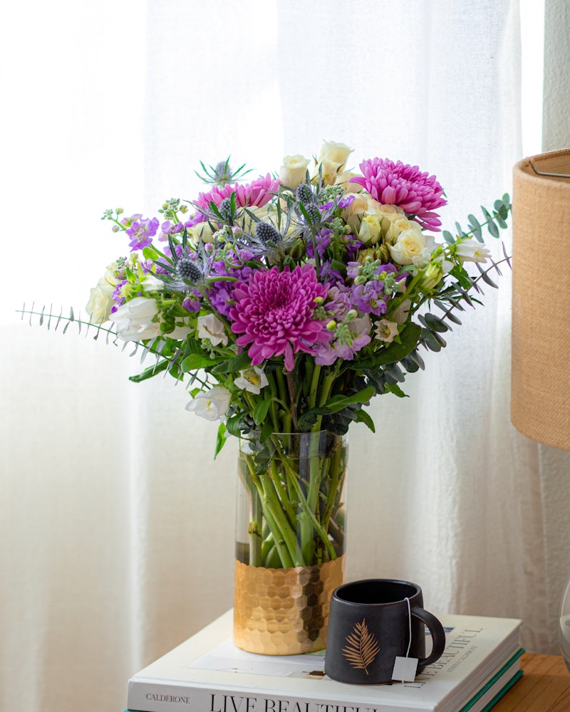 A vibrant bouquet of pink, purple, and white flowers in a gold-accented vase on a table with books and a black coffee mug, by a sheer-curtained window with natural light.