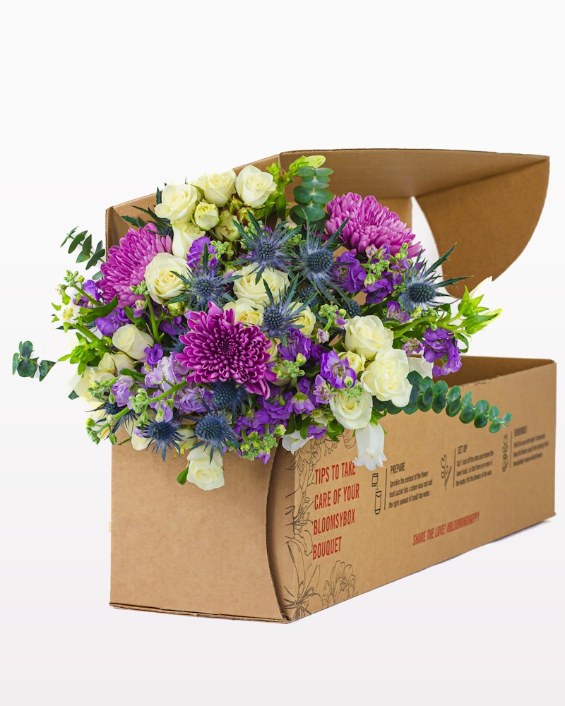 A vibrant floral arrangement featuring purple chrysanthemums, white roses, and green foliage partially inside a cardboard delivery box with care instructions.