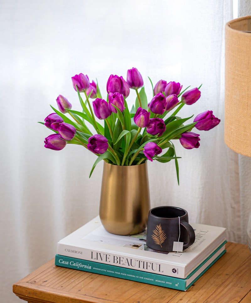 A vibrant bouquet of purple tulips in a golden vase placed on top of stylish books next to a black coffee cup, all set against a white curtain backdrop.