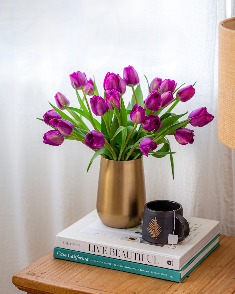 A vibrant bouquet of purple tulips in a golden vase placed on top of stylish books next to a black coffee cup, all set against a white curtain backdrop.