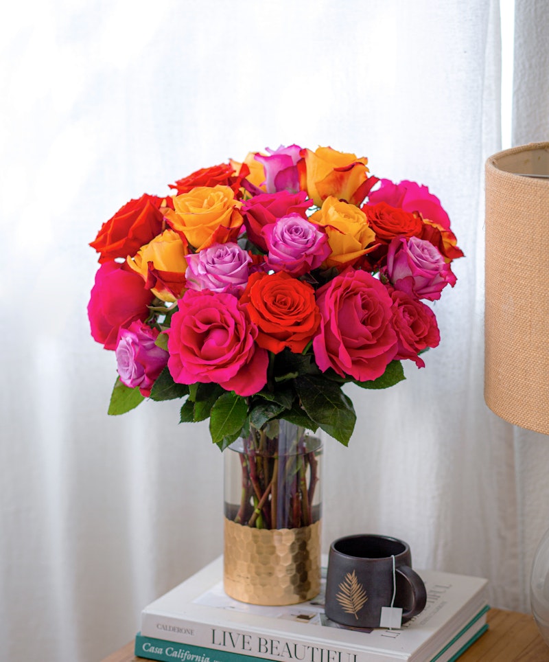 A vibrant bouquet of roses in shades of pink, orange, and yellow arranged in a clear vase on a stack of books beside a leaf-patterned mug on a wooden table.