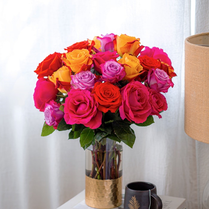 A vibrant bouquet of roses in shades of pink, orange, and yellow arranged in a clear vase on a stack of books beside a leaf-patterned mug on a wooden table.