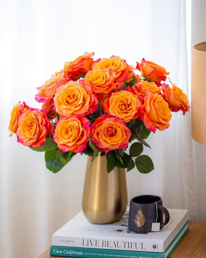 Vibrant bouquet of orange-pink roses in a gold vase on a stack of books next to a black coffee mug, with a beige lampshade in a bright room.