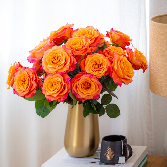 Vibrant bouquet of orange-pink roses in a gold vase on a stack of books next to a black coffee mug, with a beige lampshade in a bright room.