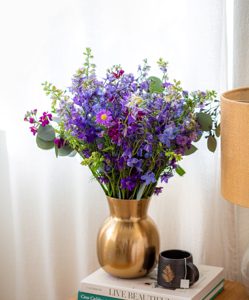 A vibrant bouquet of blue and purple flowers in a golden vase, placed on a stack of books next to a window, adding a touch of natural beauty to the cozy room decor.