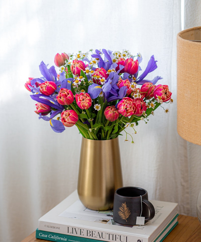 A vibrant bouquet of pink tulips with purple and white flowers in a golden vase, placed on books beside a coffee cup on a table with a sheer curtain backdrop.