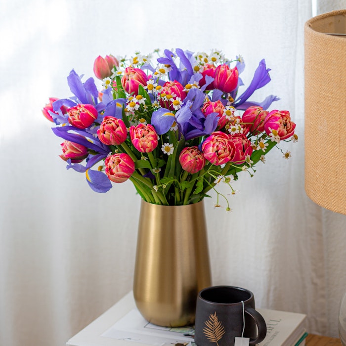 A vibrant bouquet of pink tulips with purple and white flowers in a golden vase, placed on books beside a coffee cup on a table with a sheer curtain backdrop.