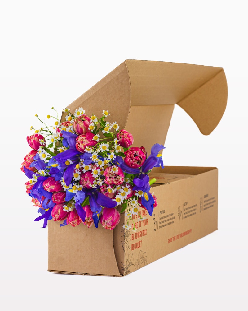 A vibrant bouquet of pink and purple flowers with green leaves and small white accents, artistically arranged and partially wrapped in a brown cardboard box.