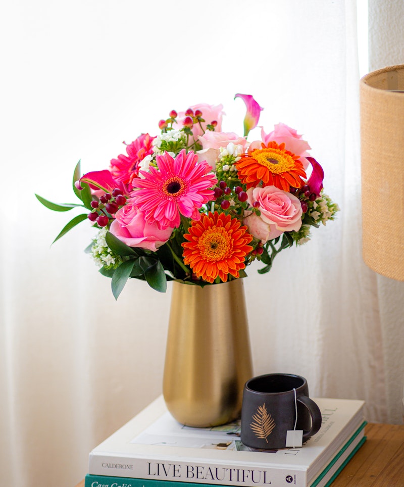 Vibrant bouquet of pink and orange flowers, including gerberas and roses, in a golden vase on a stack of books beside a black mug, with natural light.