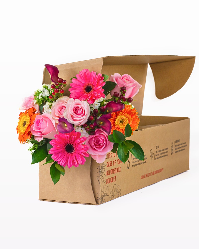 Vibrant bouquet of fresh flowers including pink gerberas, roses, and orange blooms emerging from an open cardboard box with care instructions on the side.