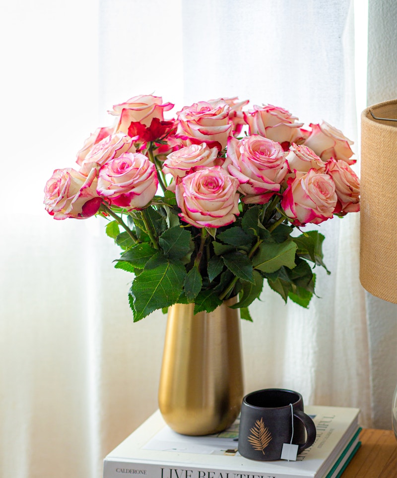 A vibrant bouquet of pink and white roses arranged in a gold vase on a side table with books and a black coffee mug, near a window with sheer curtains.