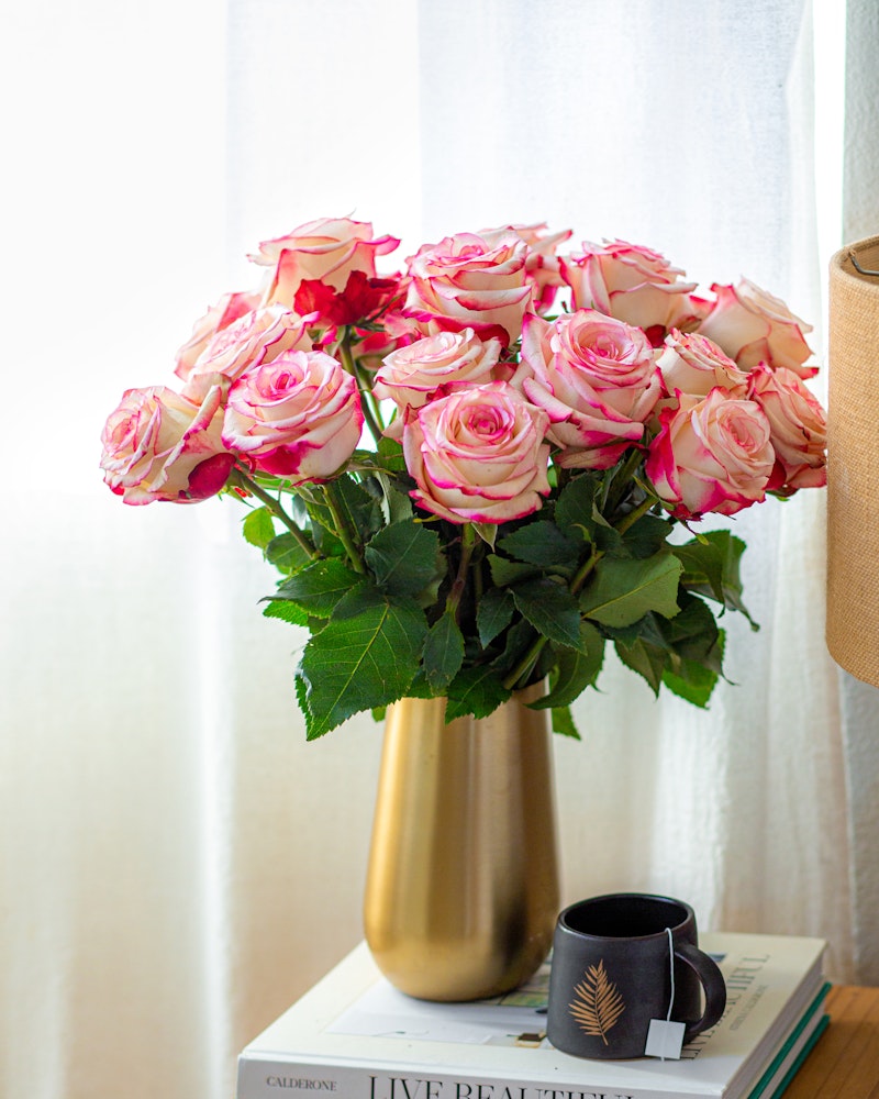 A vibrant bouquet of pink and white roses arranged in a gold vase on a side table with books and a black coffee mug, near a window with sheer curtains.