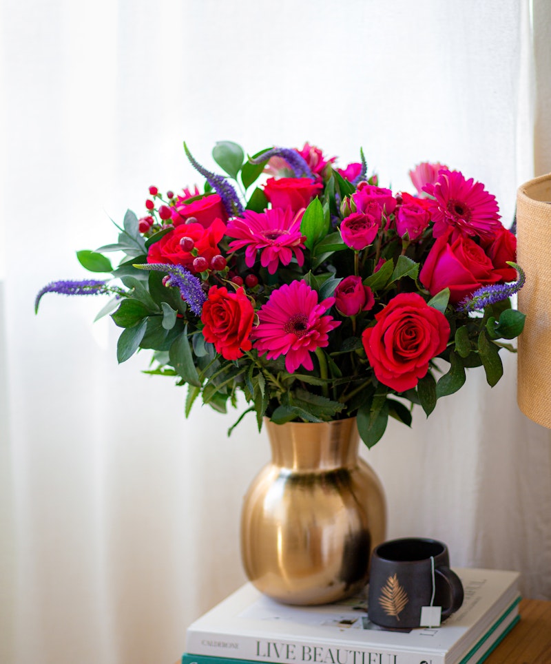 Vibrant floral arrangement with pink gerberas, red roses, and purple accents in a gold vase on a stack of books beside a window with sheer curtains.