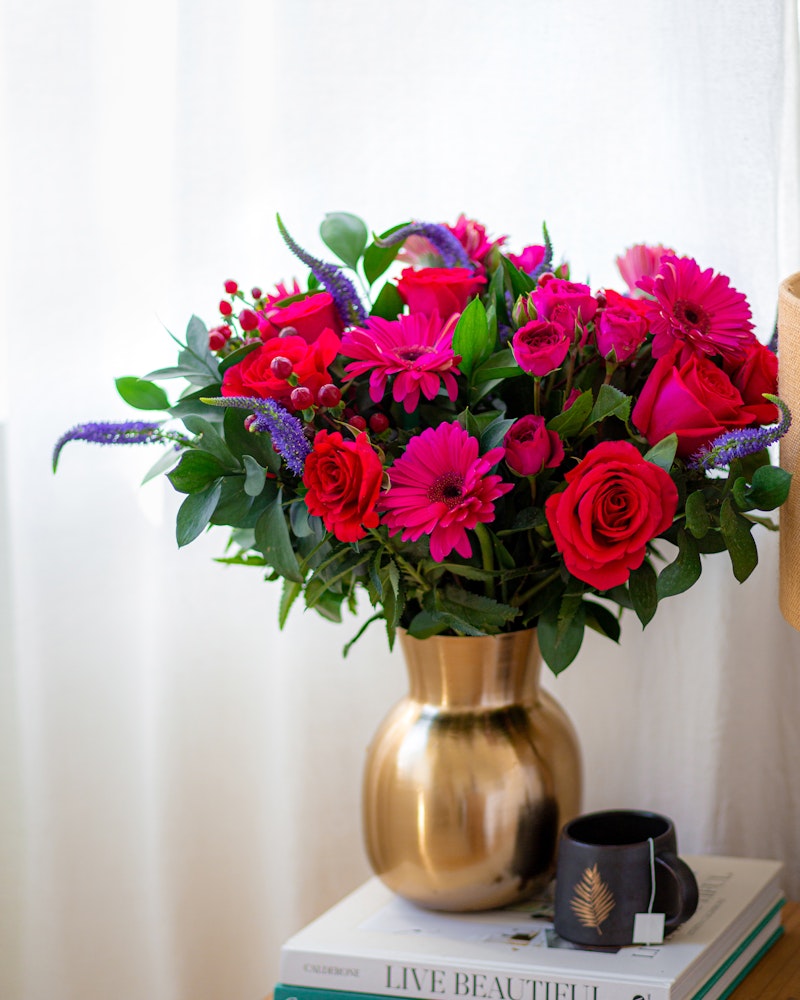 Vibrant floral arrangement with pink gerberas, red roses, and purple accents in a gold vase on a stack of books beside a window with sheer curtains.