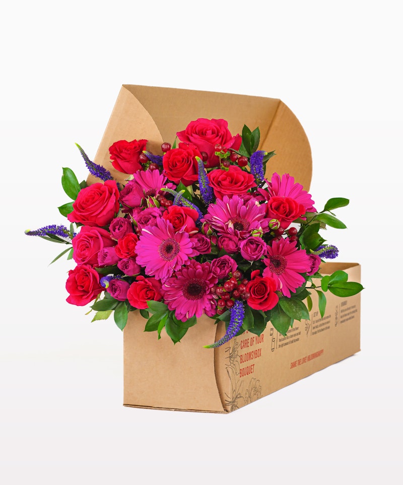 A vibrant bouquet of red roses, pink gerberas, and purple flowers neatly arranged in a cardboard flower box, showcasing a fresh and colorful floral delivery.