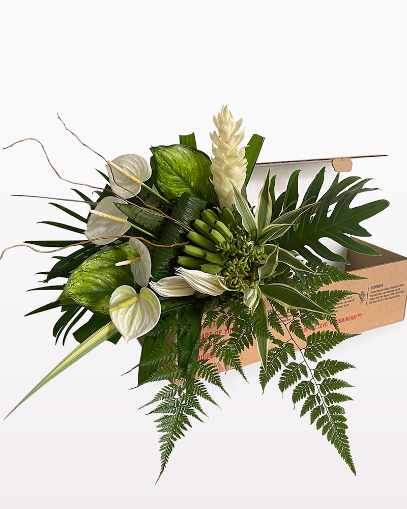 Elegant bouquet of green and white flowers with various foliage, featuring ferns and a monstera leaf, presented beside a brown cardboard box on a white background.