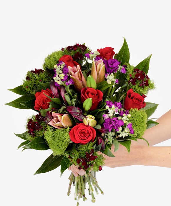 Bouquet of flowers with red roses, pink lilies, purple accents, and green foliage held by a person against a white background, ideal for special occasions.