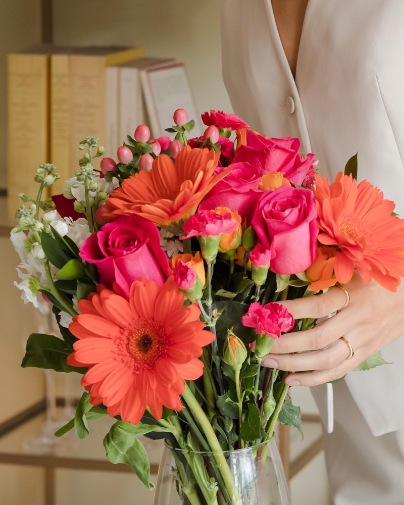 A woman in a white outfit holding a vibrant bouquet featuring orange gerberas, pink roses, and assorted greenery with a blurred background.