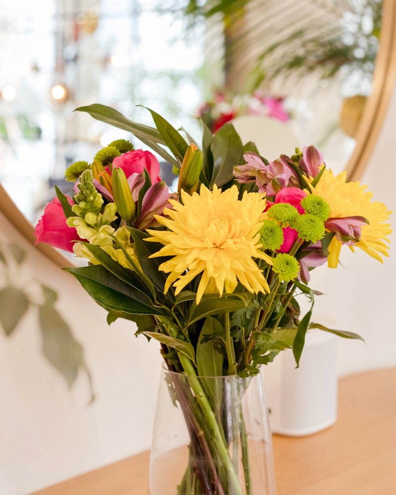 Vibrant bouquet of fresh flowers with prominent yellow dahlias and green accents in a clear vase on a wooden table, reflecting in a round mirror in a bright room.