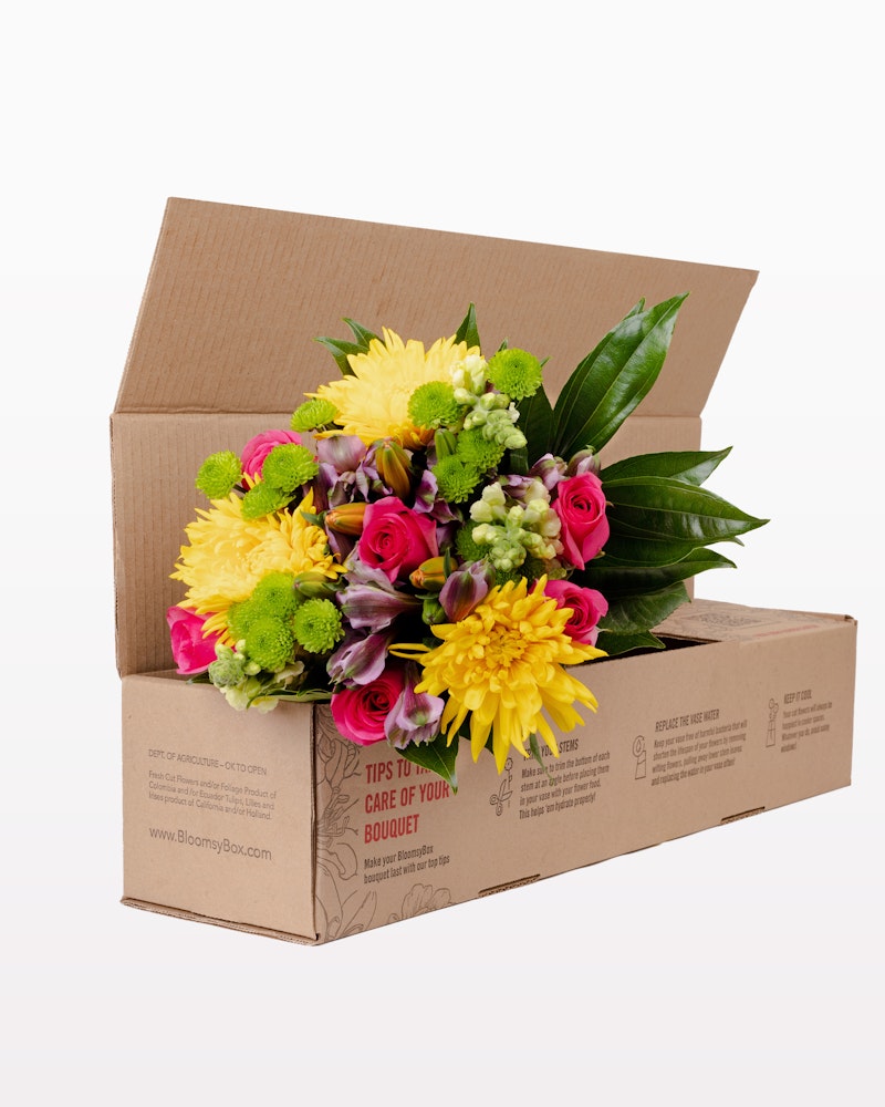 Vibrant bouquet of fresh flowers including yellow, pink, and green blooms with lush foliage, packaged neatly in an open cardboard box against a white background.