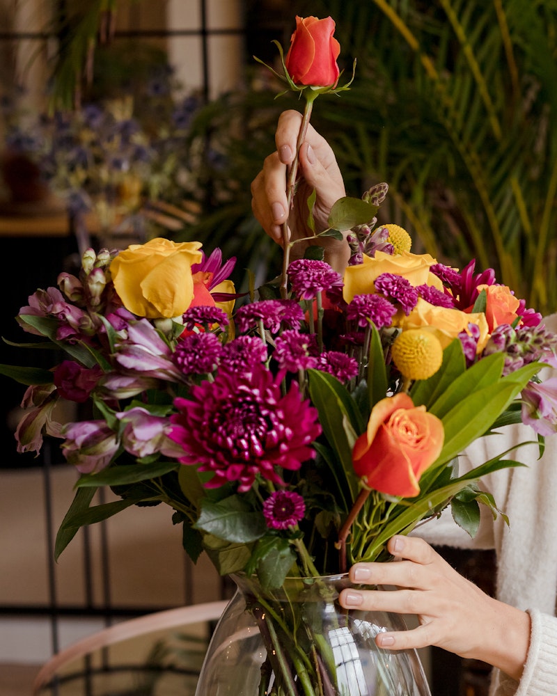 Close-up of a person's hands arranging a colorful bouquet with orange roses, purple chrysanthemums, and assorted flowers in a clear vase against a plant-filled background.