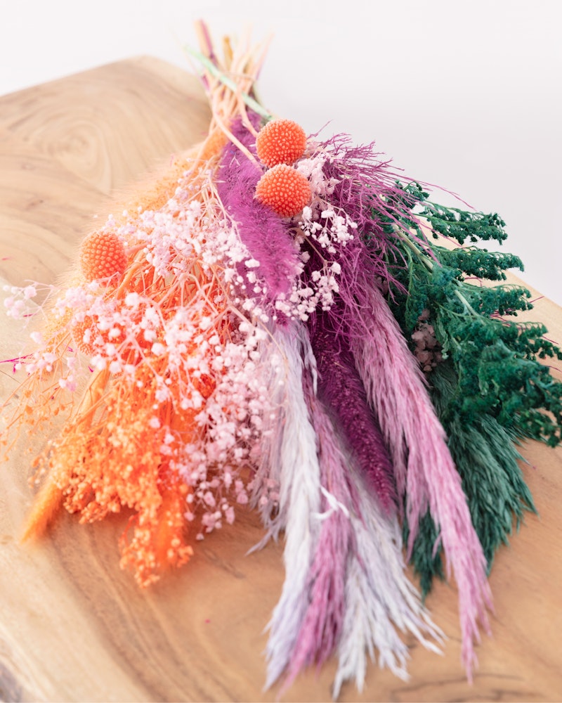 Colorful dried flower bouquet featuring vibrant shades of orange, purple, and green with a mix of textures on a wooden surface against a white background.