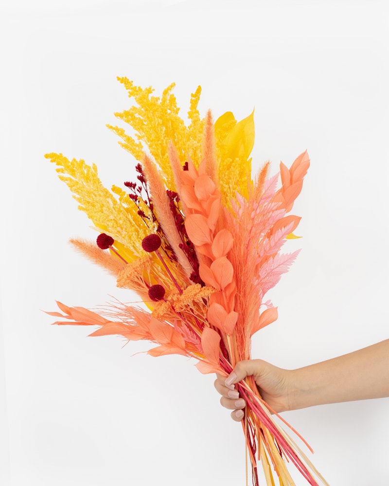 A hand holding a vibrant bouquet of artificial flowers in shades of yellow, orange, and pink against a white background, perfect for festive home decor.