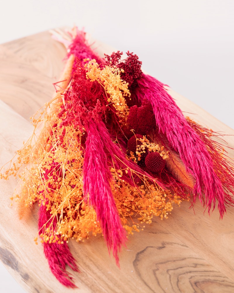 Vibrant arrangement of dried flowers showcasing deep pink and bright yellow hues on a natural wooden board with a soft white background, demonstrating home decor elegance.