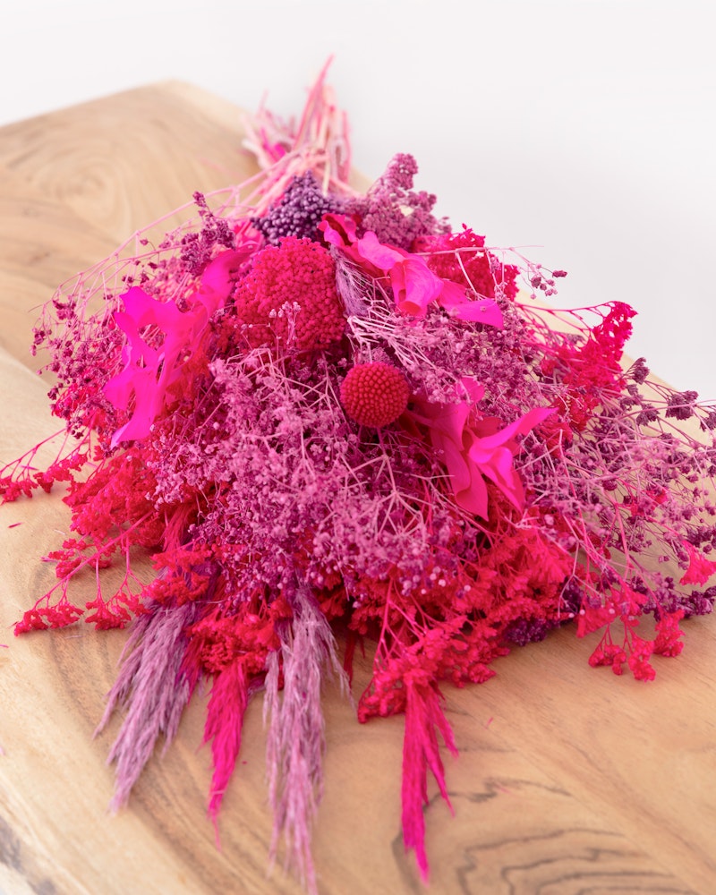 Vibrant bouquet of fuchsia dried flowers with a variety of textures, prominently featuring pink grasses and spherical blooms, artistically arranged on a wooden surface.