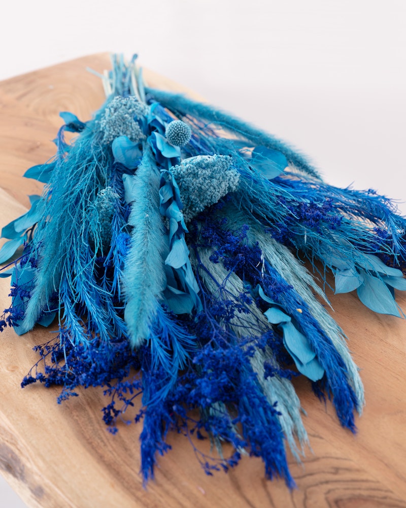 Blue and teal dried flower arrangement featuring a mix of feathers and textured botanical elements on a wooden surface, displaying a vibrant contrast of colors.