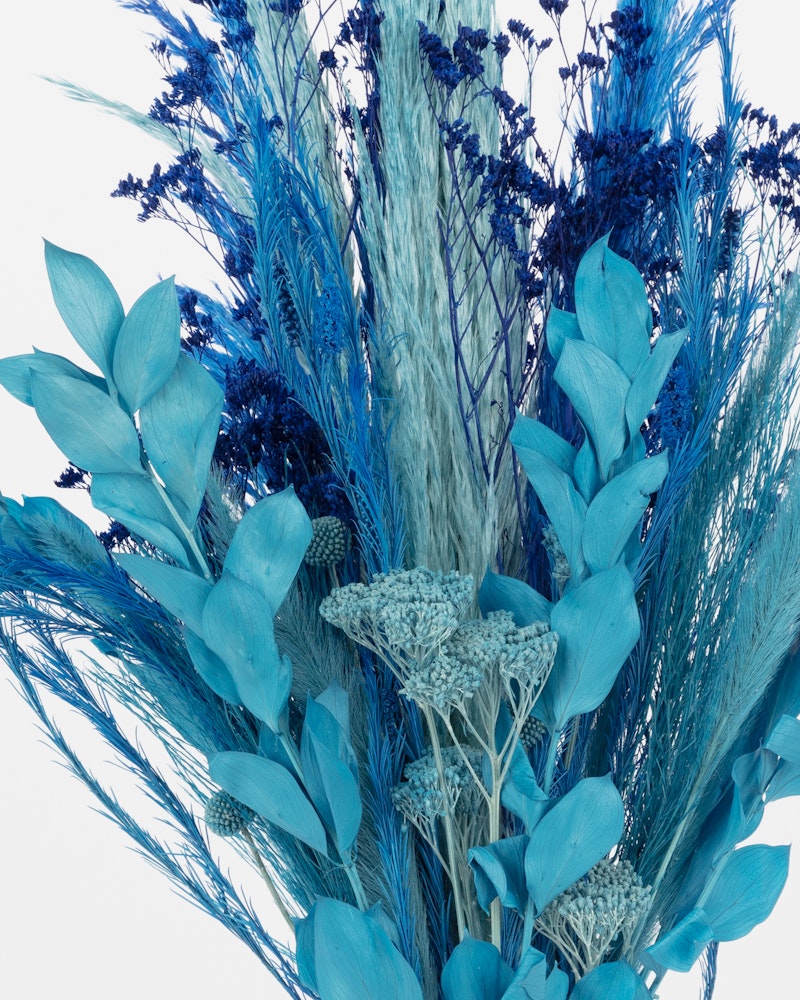 Vibrant blue artificial flowers and plants arrangement featuring various textures and tones with pine cones against a white background.