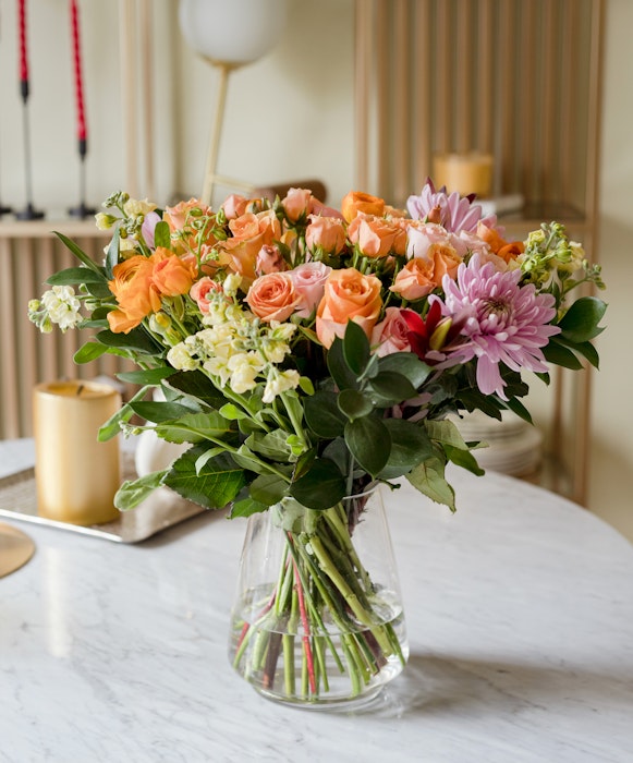 A beautiful bouquet of mixed flowers, including orange roses and pink lilies, arranged in a clear glass vase on a marble table with elegant home decor in the background.