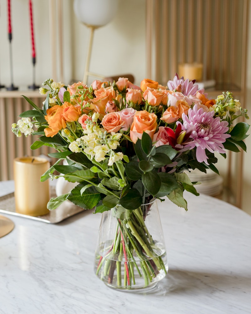 A beautiful bouquet of mixed flowers, including orange roses and pink lilies, arranged in a clear glass vase on a marble table with elegant home decor in the background.