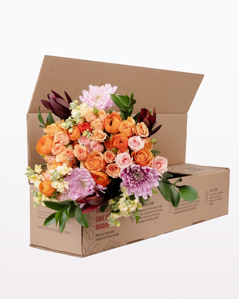 A vibrant bouquet of fresh flowers including orange roses, purple lilies, and pink blooms arranged beautifully in an open brown cardboard box with the text "bloomsybox.com".