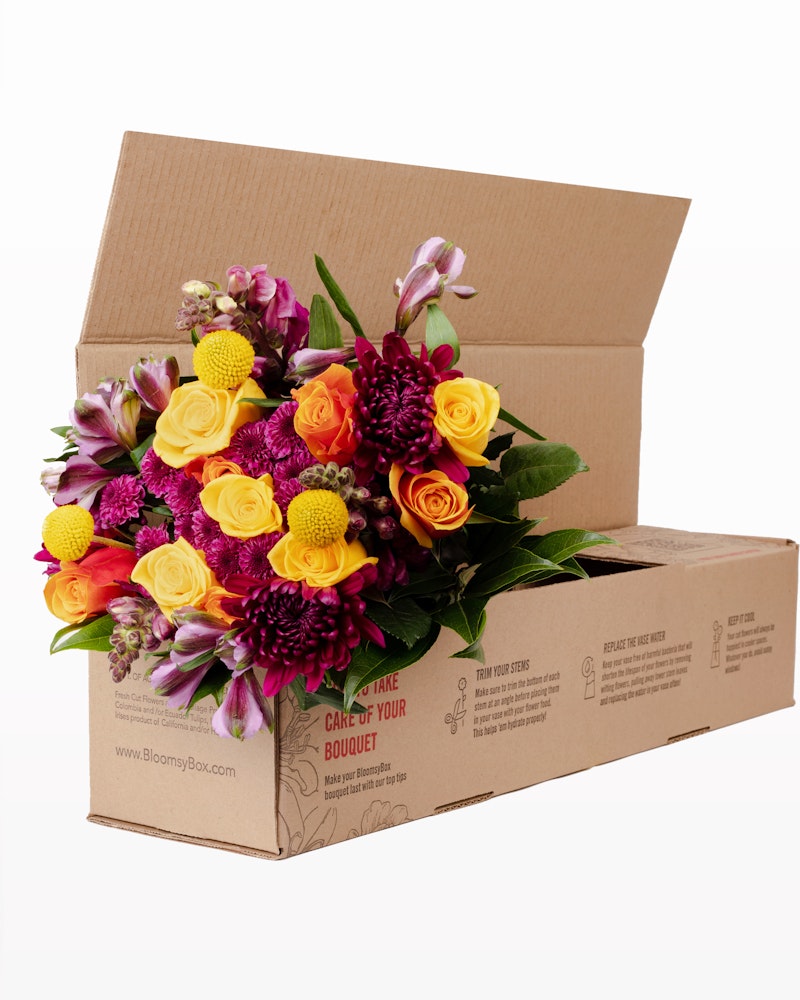 A vibrant bouquet of flowers, featuring purple, pink, and yellow blooms, creatively arranged in an open cardboard box with care instructions visible on the side.