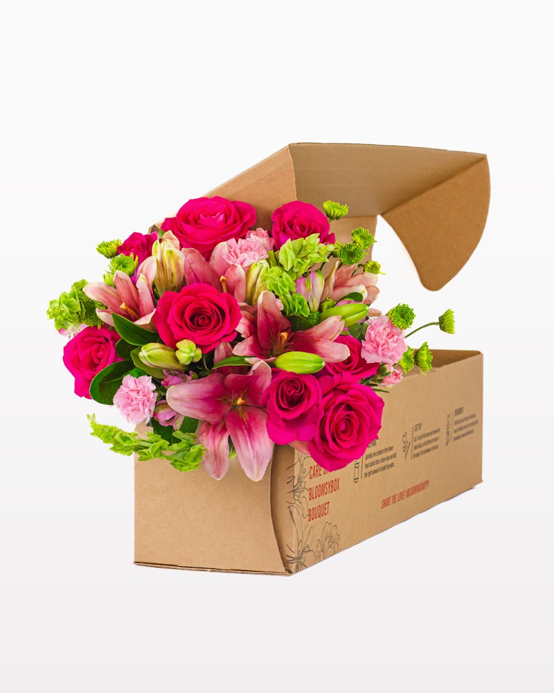 A vibrant bouquet of pink roses and alstroemeria neatly arranged in a partially opened, eco-friendly brown cardboard box on a white background.