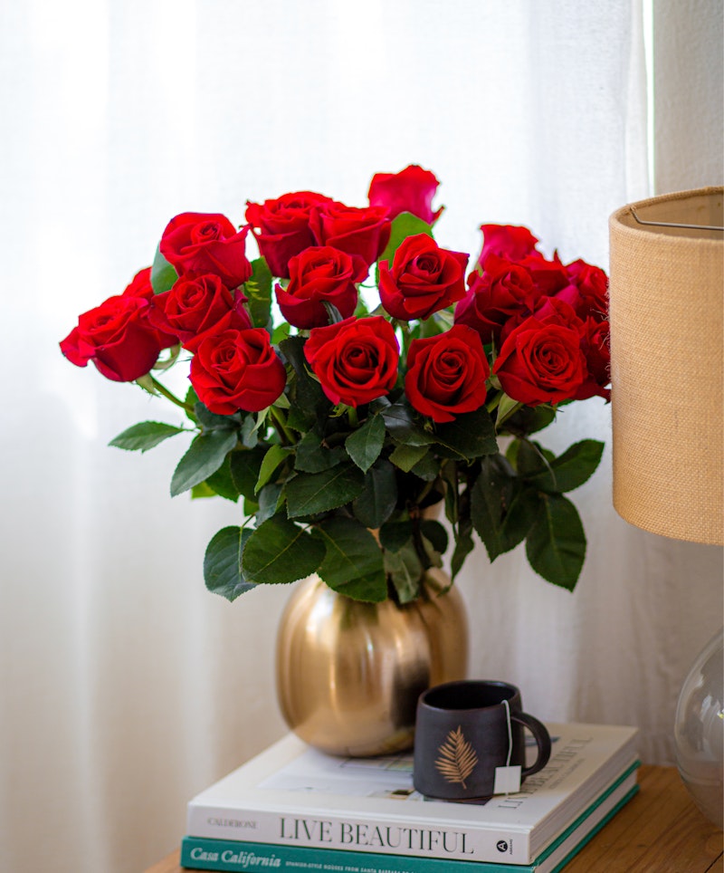 Vibrant bouquet of red roses in a golden vase on a stack of books beside a window, with a small coffee cup on a white tablecloth, conveying a cozy home atmosphere.