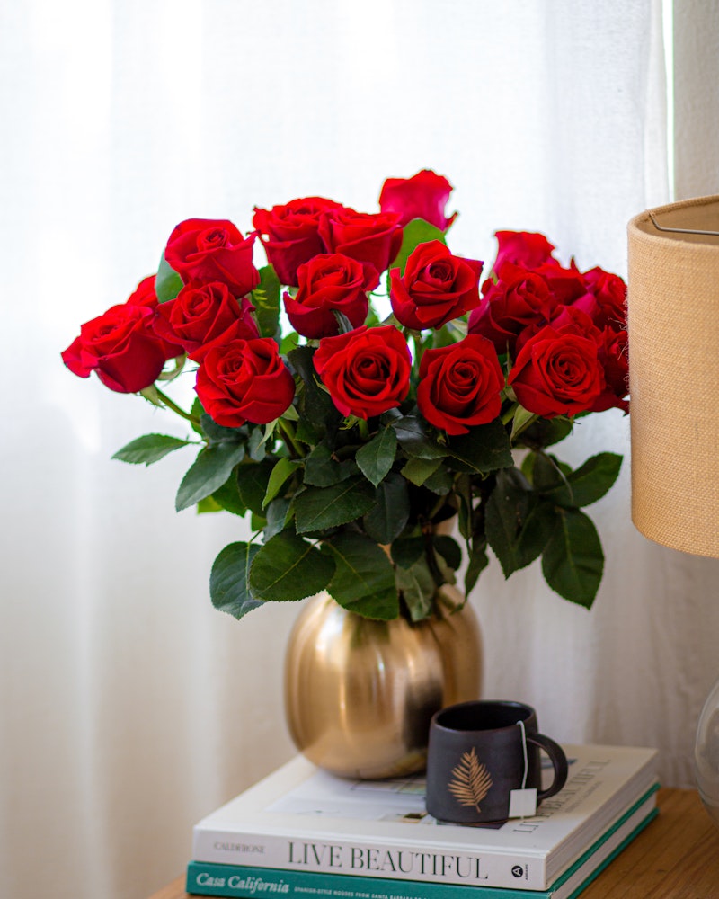 Vibrant bouquet of red roses in a golden vase on a stack of books beside a window, with a small coffee cup on a white tablecloth, conveying a cozy home atmosphere.