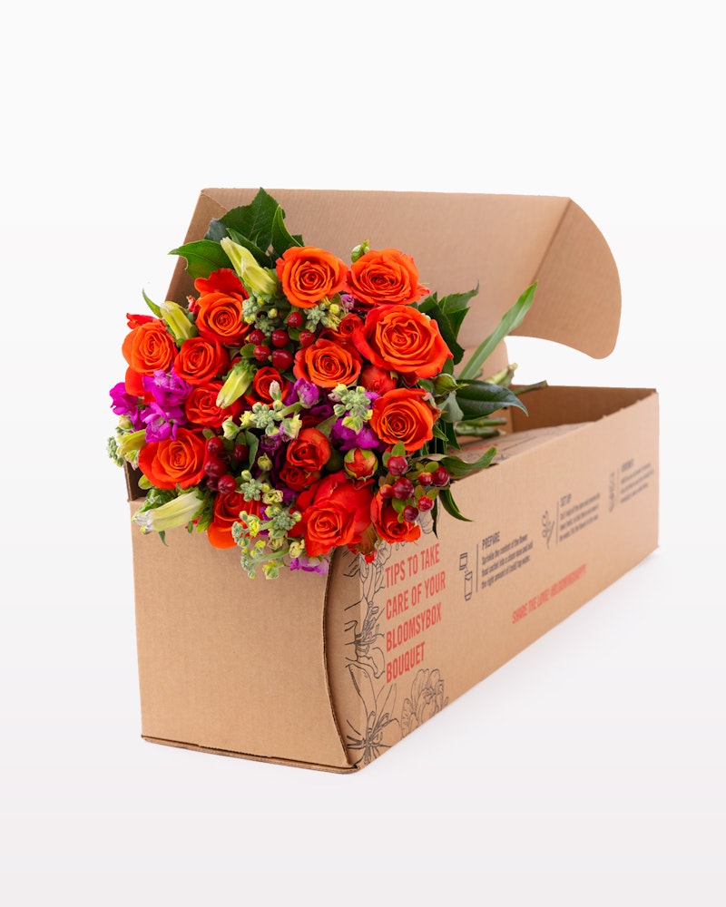Vibrant bouquet of orange roses and purple flowers packaged in a cardboard box with care instructions printed on the side, ready for delivery against a white background.