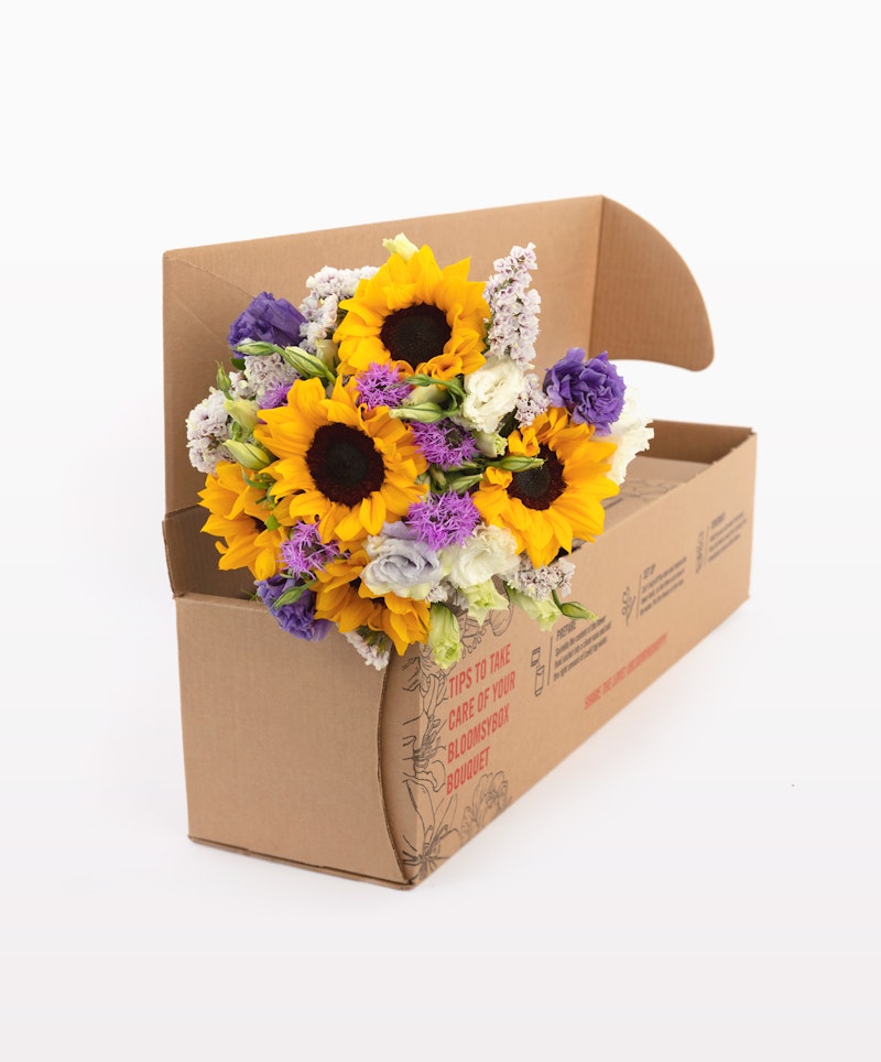 Bouquet of sunflowers and purple flowers presented in a cardboard delivery box against a white background, showcasing a fresh floral arrangement ready for shipment.