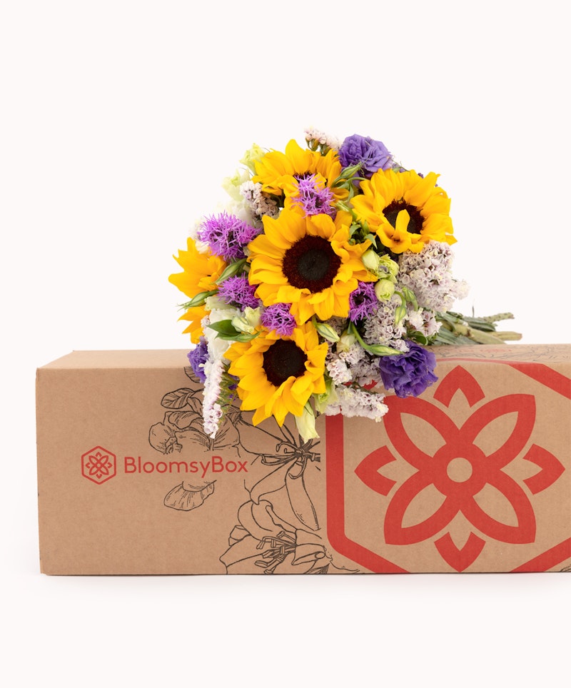 Vibrant bouquet of sunflowers and assorted flowers spilling from a BloomsyBox, showcasing a fresh floral delivery service with a branded cardboard box on a white background.