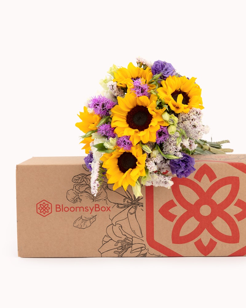 Vibrant bouquet of sunflowers and assorted flowers spilling from a BloomsyBox, showcasing a fresh floral delivery service with a branded cardboard box on a white background.