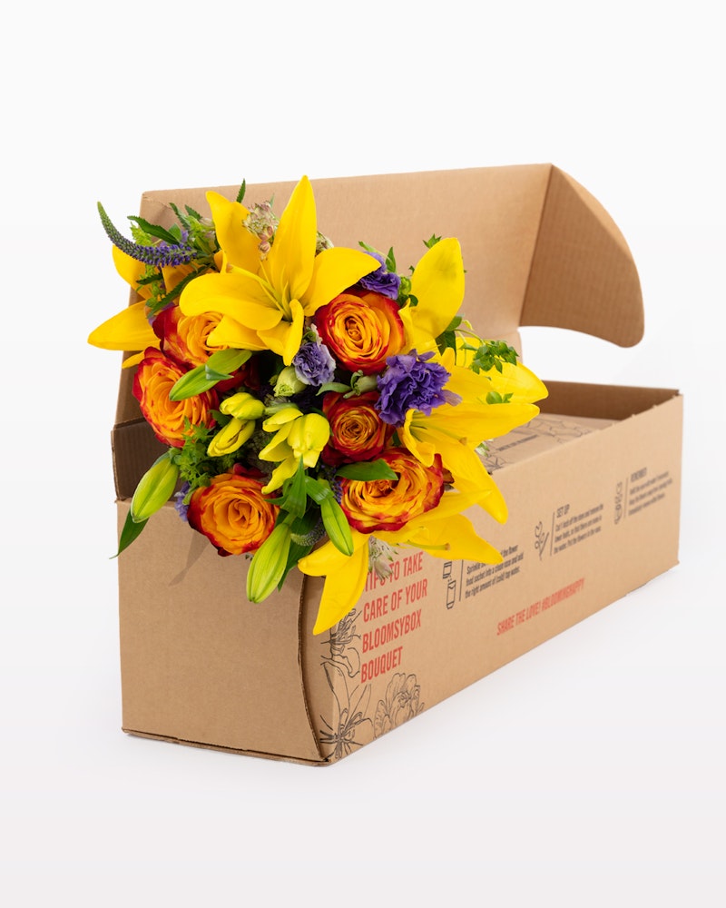Vibrant yellow and purple flower bouquet with lilies, roses, and greenery emerging from an open cardboard box against a white background, highlighting fresh flower delivery service.